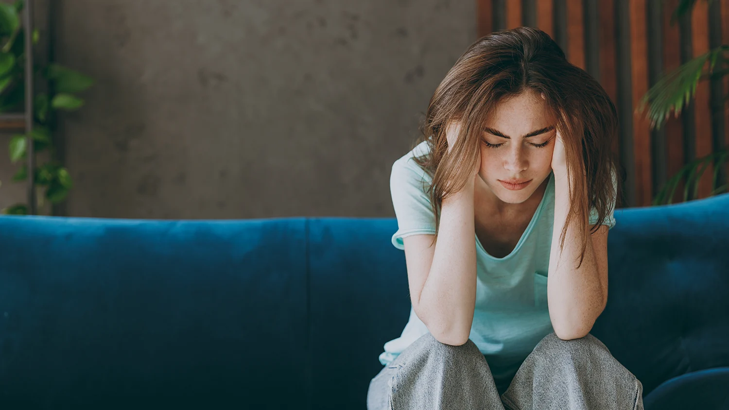 A woman sits on a couch and holds her head from headache pain
